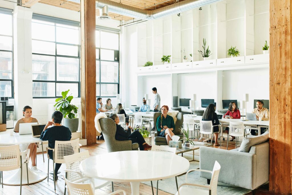 Open plan office with high ceilings, big windows and a mix of different types of spaces. Two people sitting at a table with laptops facing each other. A few feet away two people in armchairs chatting beside a coffee table. A few feet beyond them, three people with devices sitting at a table working together. Behind them against the wall a row of individual workspaces. In the corner another group of five work together. A few plants populate the mostly neutral coloured spaced.