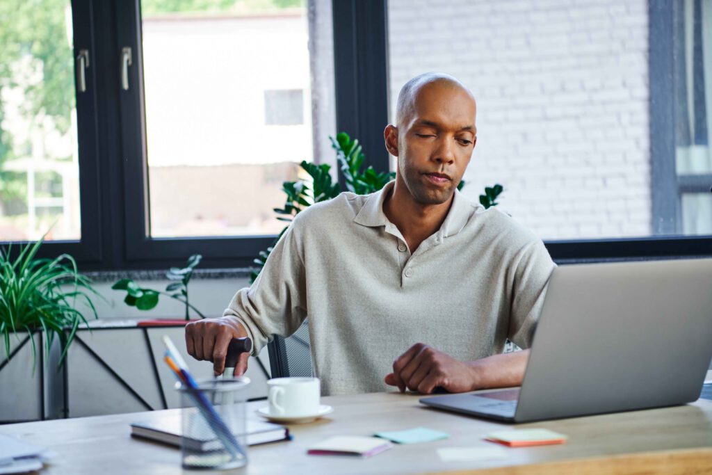 A light skinned black man with a bald head sits at a desk looking at a laptop. He has myasthenia gravis which has weakened his muscles. His face droops slightly, one eye appears to be closed. His right hand rests on a walking stick.